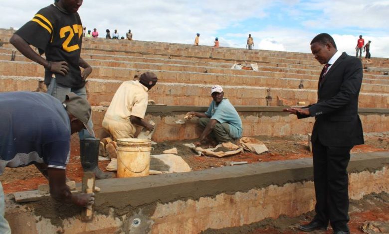 Workers are building steps or seats in a Stadium and look up to a man in a nice suit who is talking and gesticulating